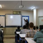 معلم در حال تدریس و نوشتن روی تخته سفید در کلاس درسی با دانش‌آموزان نشسته در میز‌های درسی