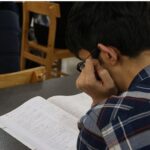 دانش‌آموز در حال مطالعه و نوشتن تکالیف درسی در میز کلاس درس با صندلی چوبی