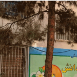 نمای بیرونی ساختمان مدرسه با تابلوی رسمی، درب‌های فلزی، پنجره‌های سفید و درختی بزرگ در جلو