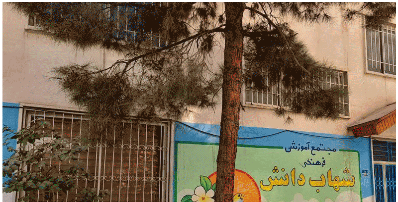 نمای بیرونی ساختمان مدرسه با تابلوی رسمی، درب‌های فلزی، پنجره‌های سفید و درختی بزرگ در جلو