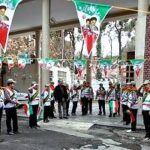 دانش‌آموزان مدرسه در حیاط مدرسه ایستاده‌اند و در یک مراسم سرود جمعی شرکت می‌کنند، پرچم‌های رنگی ایرانی از بالا آویزان است