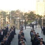 دانش‌آموزان مدرسه شهید مطهری در حیاط ایستاده‌اند در صفبندی صبح با ساختمان‌های مدرسه در پس‌زمینه