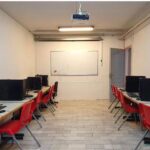 نمای کلی اتاق کامپیوتری مدرسه با کامپیوترهای رومیزی، صندلی‌های قرمز، میز‌های کار و تخته سفید