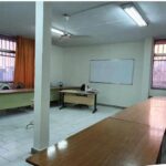 نمای درونی کلاس درس خالی با میز‌های چوبی، صندلی‌های آبی، تخته‌سفید، ستون‌های بتنی سفید، نور طبیعی از پنجره‌های بزرگ و نور مصنوعی سقفی