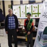 عکس از یک مراسم رسمی مدرسه با حضور کارکنان و دانش‌آموزان در کنار بنری که متن‌های تجلیل روی آن نوشته‌شده است