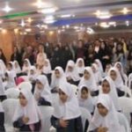 گروهی از دختران دانش‌آموز با حجاب سفید در یک مراسم یا جشن در سالن داخلی روشن با فانوس‌های نور سفید