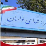 تابلوی آبی نصب‌شده‌ی درب ورودی مدرسه راهنمایی شهدای لواسان با نام مدرسه به خط فارسی و پرچم ایران در بالای تابلو