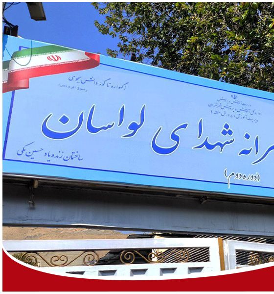 تابلوی آبی نصب‌شده‌ی درب ورودی مدرسه راهنمایی شهدای لواسان با نام مدرسه به خط فارسی و پرچم ایران در بالای تابلو