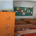 کلاس درس مجهز به میزهای قرمز و کمدهای نارنجی، با تابلوی اطلاع‌رسانی سبز رنگ حاوی کارهای هنری دانش‌آموزان