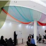 سالن داخلی مدرسه آماده‌شده برای برگزاری مراسم و رویداد، با سقف تزئین‌شده به رنگ‌های قرمز و آبی و صورتی، و گروهی از شرکت‌کنندگان