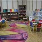 اتاق یادگیری کودکان با صندلی‌های رنگی، قالی‌های رنگین و چند رنگ، و قفسه‌های سفید مملو از کتاب‌ها و اسباب‌بازی‌های آموزشی