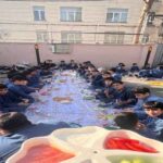 دانش‌آموزان مدرسه در حیاط نشسته در حالی که در یک پروژه هنری و خلاقانه گروهی با نقاشی شرکت می‌کنند.