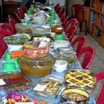 میز جشن و مهمانی با انواع غذا، میوه، نوشیدنی و دسر در کنار کتابخانه‌ای با صندلی‌های قرمز رنگ