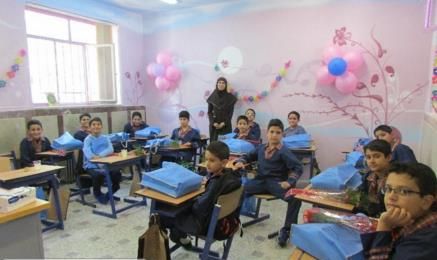 نمای داخلی کلاس درسی تزیین‌شده با بادکنک و بنرهایی رنگی، دانش‌آموزان بر روی صندلی‌های آبی نشسته‌اند و یک معلم ایستاده در کنار آنها