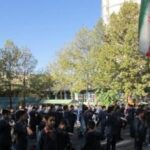 گروهی بزرگ از دانش‌آموزان و کارکنان مدرسه در حیاط ایستاده‌اند، پرچم ایران بر فراز ساختمان مدرسه به نمایش درآمده است