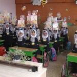 کلاسی مع دانش‌آموزان ایستاده در کنار میزهای سبز و تزیینات کاغذی رنگی روی دیوار