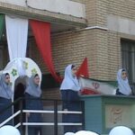 دانش‌آموزان دختر با لباس فرم و حجاب سفید در حال شرکت در یک مراسم رسمی در محوطه‌ی ساختمان مدرسه، با پرچم‌های ملی و بنرهای رنگی