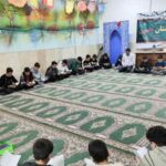 دانش‌آموزان نشسته بر روی موکت سبز نماز در نمازخانه مدرسه، حین فعالیت مطالعه و نماز با تزئینات اسلامی دیواری