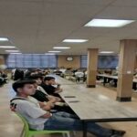 دانش‌آموزان در حال شرکت در یک جلسه‌ی اجتماعی یا فعالیت گروهی در سالن بزرگ مدرسه، نشسته‌اند روی صندلی‌های سبز و میزهای طویل