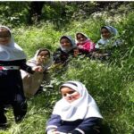 گروهی از دانشآموزان دختر با لباس فرم سفید و سرمهای در حیاط مدرسه در میان گیاهان سبز