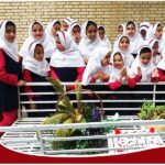 دانشآموزان دختر پوشیده به لباس مدرسه (روپوش قرمز و روسری سفید) در حال شرکت در رویداد باغبانی، ایستاده دور یک باغچه مزیّن با گلهای رنگی، گیاهان و تاج طلایی