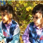 دو دانشآموز در حیاط مدرسه، بین گیاهان سبز و درختان پر برگ در یک روز آفتابی