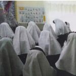 گروهی از دانشآموزان دختر با لباس مدرسه نشستهاند، یک معلم یا مربی ایستاده است، تخته سیاه و تابلوی آموزشی در پسزمینه قابلرویت است