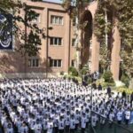 تجمع گروهی صدها دانش‌آموز با لباس یکنواخت در حیاط مدرسه؛ ساختمان آجری و درختان در پس‌زمینه