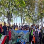 گروهی از دانش‌آموزان و معلمان در حیاط مدرسه برای یک مراسم گردهمایی جمعی، نگه‌دارنده بنری با نوشته فارسی و پرچم ایران