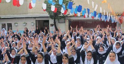 تجمع جمعی دانش‌آموزان با لباس یکدست و حجاب سفید در حیاط مدرسه با بالن‌های رنگی و نوارهای تزیینی