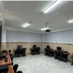 نمای سایت کامپیوتری مدرسه با چندین رایانه دسک‌تاپ و صندلی‌های سیاه روی میز‌های چوبی