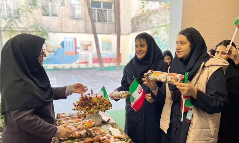 دانش‌آموزان در حال شرکت در یک رویداد فرهنگی و غذایی سنتی ایرانی در فضای باز مدرسه؛ برخی با پرچم ایران و دستفروشی غذاهای سنتی.