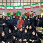 گروهی از دانش‌آموزان دختر به همراه یک معلم یا مهمان در مراسم جشن مدرسه، میان بادکنک‌های رنگی و پرچم‌های ایرانی، در حال عکس‌گرفتن با غذاهای ایرانی