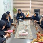 گروهی از دانش‌آموزان در حال برگزاری جلسه یا نشست تیمی دور یک میز در اتاق جلسات مدرسه، با حضور یک مربی یا معلم