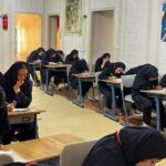 دانش‌آموزان دختر در حال انجام کار نوشتاری و امتحان در کلاس درس، با معلم ایستاده در پس‌زمینه