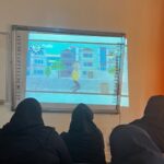 دانش‌آموزان دختر در حال تماشای درس تعاملی روی صفحه‌نمایش سفید‌برد در کلاس درس با تخته‌سیاه و پنجره‌های روشن