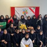 گروهی از دانش‌آموزان دختر در حال شرکت در یک رویداد مدرسه‌ای، با پوستر و بنری در پس‌زمینه