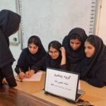 دانش‌آموزان دختر در حال انجام یک فعالیت گروهی تحت راهنمایی معلم، نشسته‌اند دور میز در کلاس درس