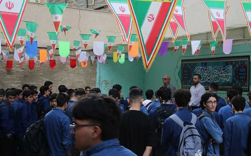 تجمع دسته‌ای از دانش‌آموزان پوشیدۀ لباس یکسان آبی در یک اتاق مدرسه‌ای با دیوارهای سبزرنگ و تزیینات پرچم‌های رنگی و ملی‌ایران برای یک مراسم یا رویداد آموزشی