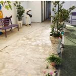 نمای حیاط مدرسه با گیاهان تزیینی، صندلی‌های رنگین، و چمن سبز
