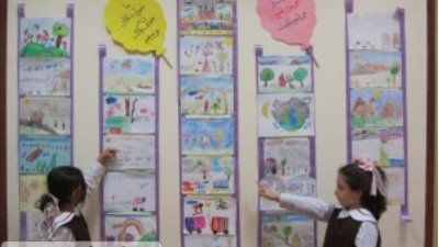 دو دانش‌آموز دبستان در حال شرکت در فعالیت هنری و نقاشی روی دیوار که پر از آثار رنگی و نقاشی‌های دانش‌آموزان است