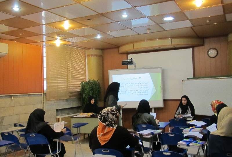 دانش‌آموزان دختر نشسته در صندلی‌های آبی در حال شرکت در یک درس، معلم در حال ارائه محتوای درسی از طریق پروژکتور در کلاسی با سقف کاذب و تجهیزات آموزشی
