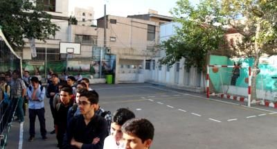 دانش‌آموزان در حیاط مدرسه ایستاده‌اند، ساختمان‌های مدرسه و فضای بیرونی دیده می‌شود