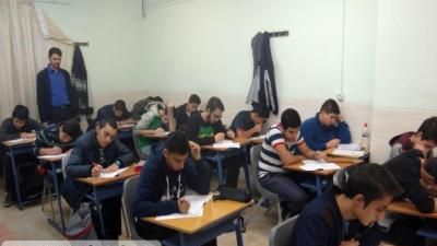 دانش‌آموزانی در حال نشستن پشت میزهای درسی در یک کلاس درس، با معلمی ایستاده در کنار تخته سیاه
