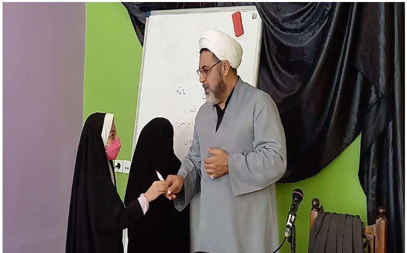معلمی در حال تقدیر از دانش‌آموزی در مراسمی درون‌مدرسه، با حضور دانش‌آموزان دیگر و میکروفون، پشت تخته‌سفید و دکوراسیون رنگی