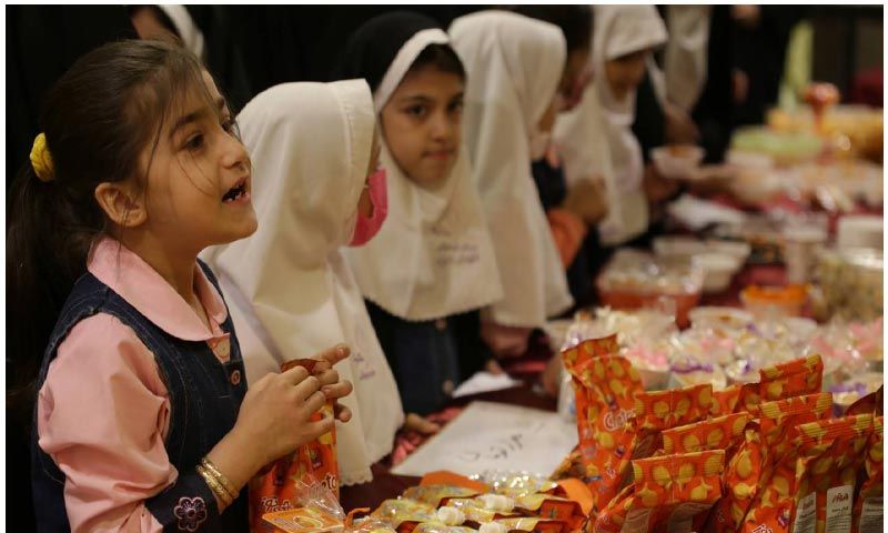 دانش‌آموزان دختر و پسر در حال انجام فعالیت هنری و ساخت گل‌های کاغذی رنگین در کنار میزهای رنگی