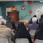 تصویری از یک جلسه سخنرانی در مدرسه که شرکت‌کنندگان نشسته‌اند و به سخنران گوش می‌دهند؛ دیوار پس‌زمینه دارای نوشتار فارسی و تزئینات است