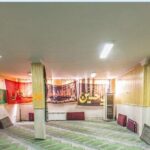 نمایی از نمازخانه مدرسه با فرش سبز، تزئینات دیواری اسلامی، و پرچم‌های سبز و قرمز