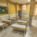 نمایش یک کلاس درس قدیمی با میزهای چوبی، صندلی‌های فلزی، تابلوی سبز و دکور داخلی از دوره تاریخی ایران