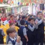 گروهی از دانش‌آموزان در حال شرکت در یک مراسم یا رویداد مدرسه‌ای در فضای باز با صندلی‌های زرد و تزئینات رنگی